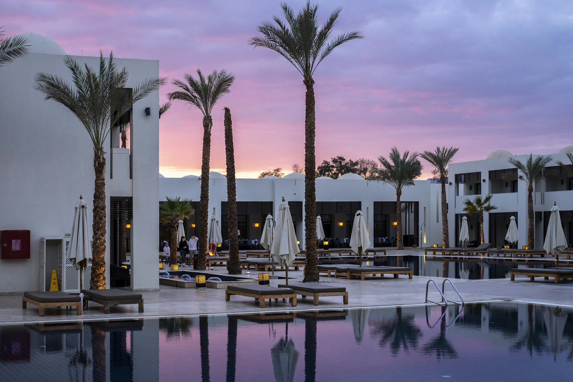 Weiße Hotelanlage in der Abenddämmerung mit Palmen, Liegen und einem Pool, in dem sich der Himmel spiegelt.
